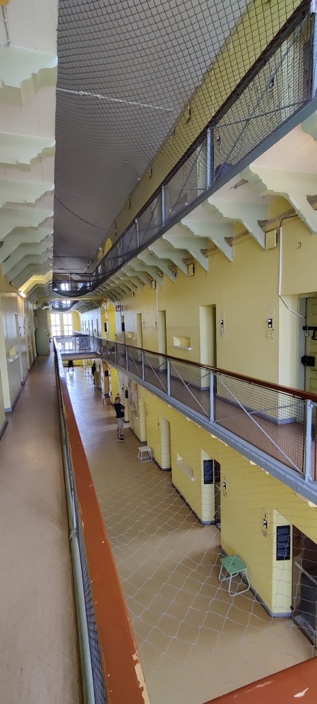 Kuva vankilan käytävästä, jossa näkyy useampi kerros samaan aikaan. Kaiteiden reunustama käytävä kiertää seinustaa. Kerrosten väliseen avoimeen tilaan on viritetty verkko, ettei alas pääse putoamaan.