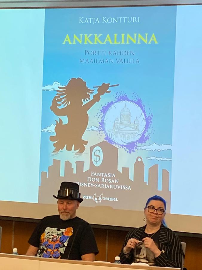 Kuva Katjasta ja hattupäisestä mieshenkilöstä pöydän takana puhumassa. Taustalla iso mainoskuva Katjan Don Rosa -kirjasta.