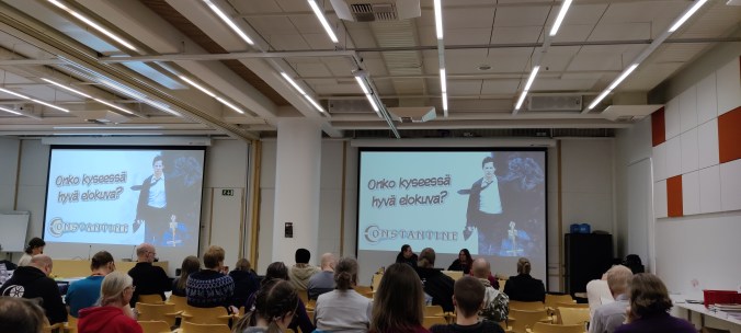 Salin takaosasta otettu kuva yleisöstä kuuntelemassa elokuva-adaptaatioesitelmää. Ruudulla kuva Constantine-elokuvasta ja kysymys, onko kyseessä hyvä elokuva.