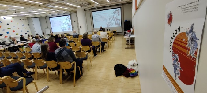 Salin takaosasta otettu valokuva yleisöstä kuuntelemassa Praedor-esitelmää. Kuvan oikealla etuosassa näkyy seinällä oleva punavalkoinen Lokacon-tapahtuman mainos.