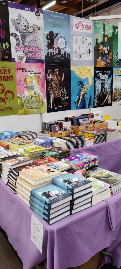 Zum Teufelin myyntipöytä, jossa etualalla pino Katjan Don Rosa -kirjoja. Taustalla sarjakuvamainosjulisteita.