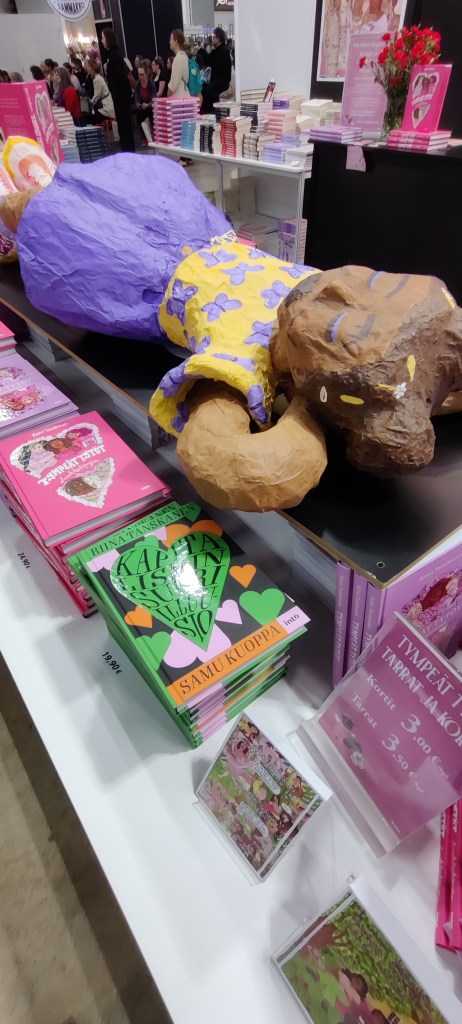 Kirjamyyntipöytä, jossa Riina Tanskasen sarjakuvia. Pöydän päällä pötköttelee paperimassasta muovailtu tympeä tyttö.