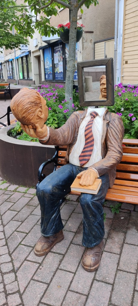 Penkillä istuva miespatsas, joka on pukeutunut farkkuihin, ruskeaan nahkatakkiin ja raidalliseen kravattiin. Patsaan toisen käden alla on kirja, toisessa tämä pitää omaa päätään. Pää katsoo ruumista, jonka pään tilalla on peili. Patsas siis katsoo itseään peilistä hämmentävällä tavalla.