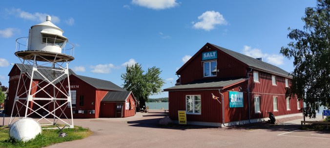 Kaksi isoa punaista puurakennusta meren rannassa. Näiden edessä seisoo valkoinen, matalan majakan näköinen rakennus. Toisen talon päädyssä lukee Museum.