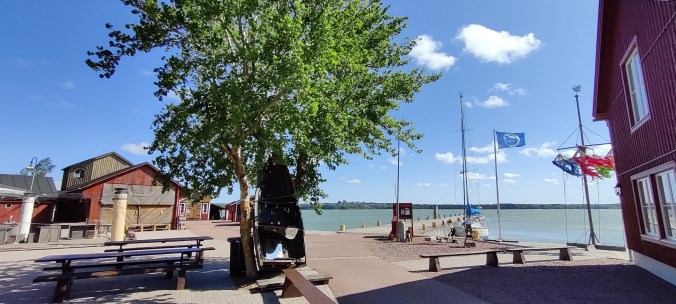 Merenranta laitureineen. Keskellä kuvaa seisoo lehtipuu, jota vasten nojaa puinen soutuvene. Taustalla on lisää punaisia rakennuksia. Oikeassa reunassa on erivärisillä nauhoilla koristeltu pylväs.