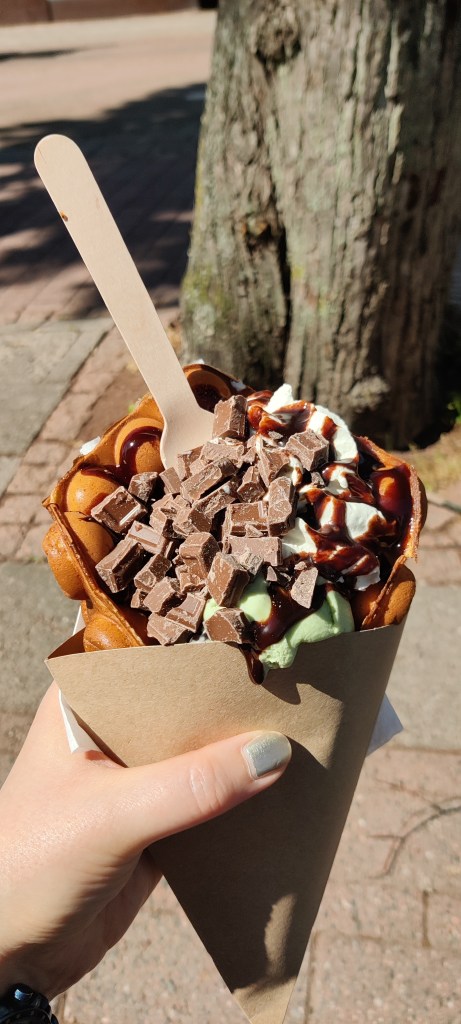 Katjan kädessä on pahvitötteröön survottu kuplavohveli, joka on täynnä jäätelöä, kermavaahtoa, suklaakastiketta ja rouhittuja suklaahippuja. Herkku syödään lusikalla.