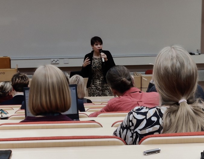 Luentosalista ihmisten päiden takaa otettu kuva Anna Kontulasta mikrofoni kädessä.