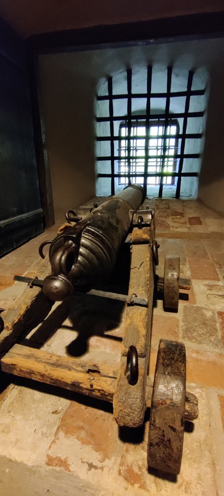 Tykki puisella jalustalla osoittaa kaltereiden suojaamaa ikkuna-aukkoa.