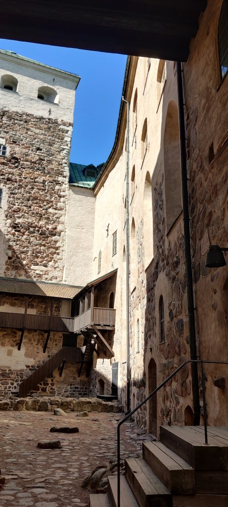 Linnan sisäpihalta otettu kuva, jossa näkyy puisia portaita ja mukulakivista pihaa. Linnan seinät ovat vaaleaa kiveä, jonka pinnassa erottuu ruskeaa kivimuuria.