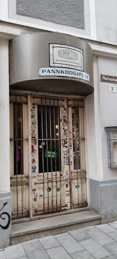Ovi, joka näyttää siltä, kuin se olisi suljettu kaltereilla. Siihen on teipattu valtavasti mainostarroja, mutta vihreä kyltti sanoo "avatud, open". Yllä olevassa kyltissä lukee Kompressor, pannkoogipubi.